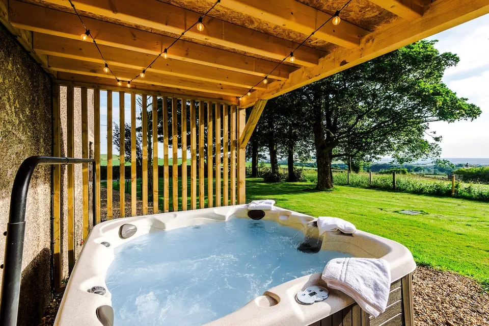 A hot tub beside a stone wall under a timber roof, with a lawn and trees beyond.