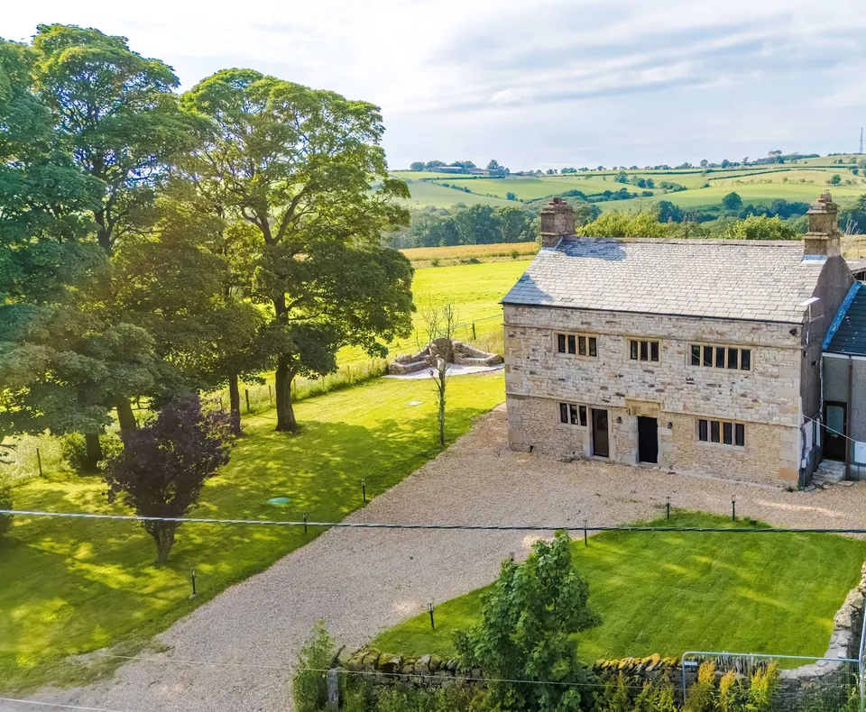 A stone house with a slate roof and two chimneys, seen from above a gravel drive, with lawns, trees and fields around it.