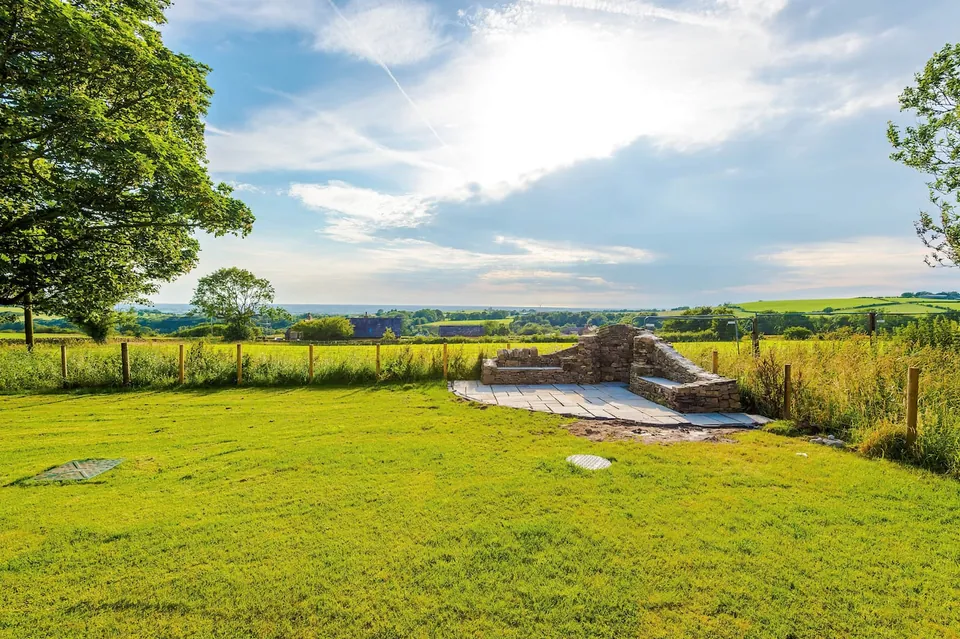 A lawn and a curved stone seat opening onto green countryside under a bright sky.