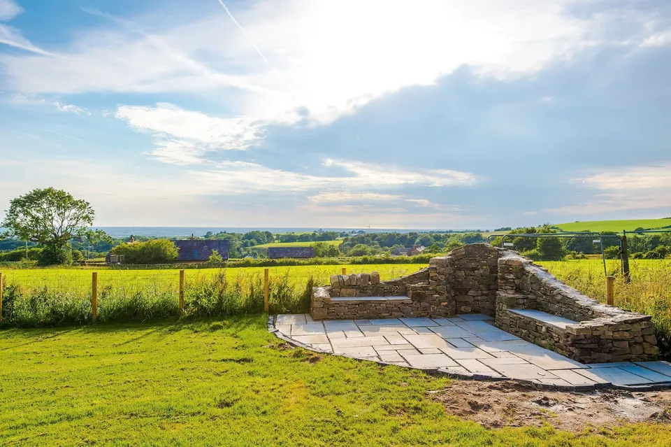 A curved stone seat on a paved lawn, looking across green and yellow fields under a blue sky.