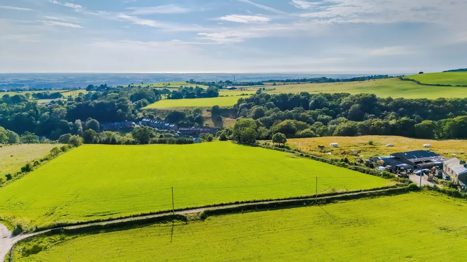 An elevated view across green fields, woodland and a small group of houses toward distant hills.
